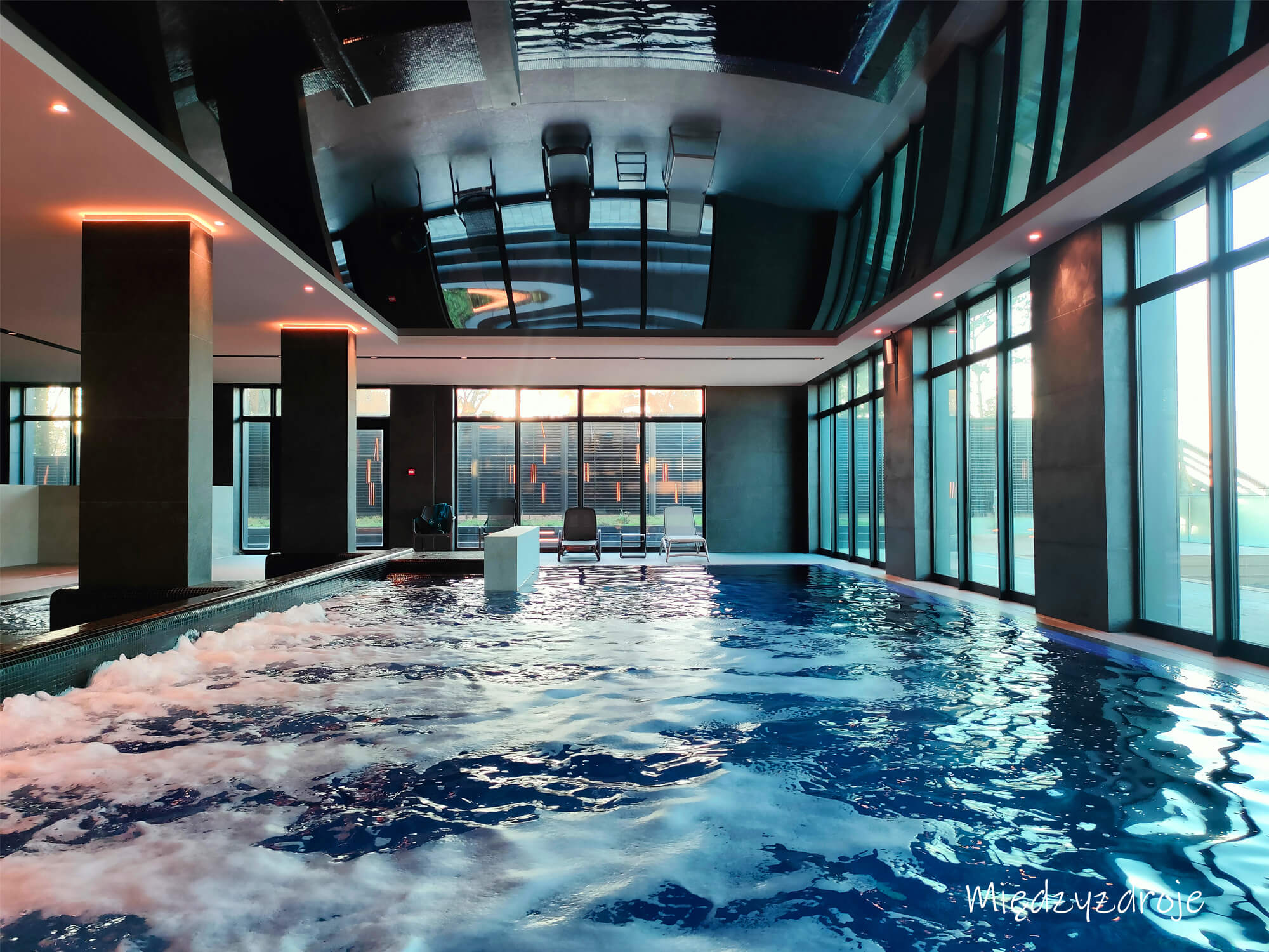Innenpool mit modernem Design, Glasfronten, Sitzliegen und spiegelnder Decke; das Wasser ist leicht aufgewühlt